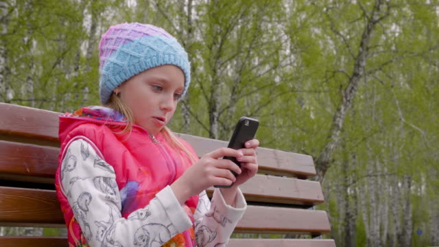 Excited-young-girl-using-phone-while-sitting-on-bench-in-park