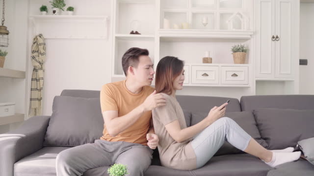Asian-couple-using-smartphone-check-social-media-in-living-room-at-home,-sweet-couple-enjoy-love-moment-while-lying-on-the-sofa-when-relax-at-home.-Lifestyle-couple-relax-at-home-concept.