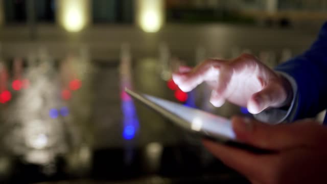 Tracking-shot-of-male-hands-swiping,-zooming-and-clicking-touchscreen-using-tablet-computer-while-man-walking-down-street-at-night