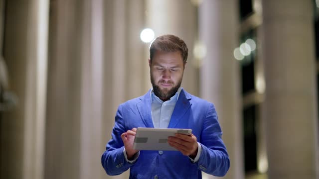 Waist-up-lockdown-shot-of-middle-adult-man-smiling-while-exchanging-messages-on-tablet-computer-standing-outdoors-in-evening