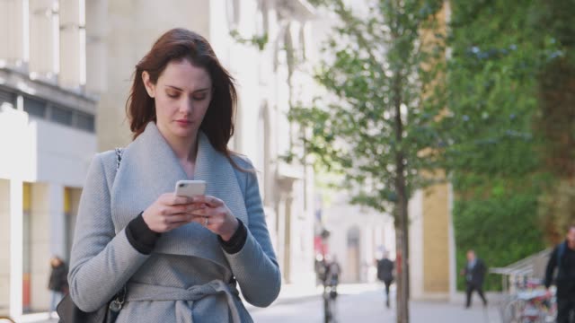 Junge-weiße-Frau-zu-Fuß-in-einer-sonnigen-London-Straße-stoppt-und-ihr-Smartphone-nutzt,-Nahaufnahme