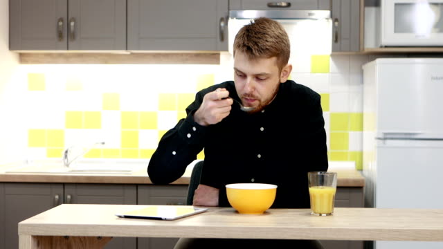 Hombre-joven-con-tableta-comiendo-el-desayuno-sentado-por-la-mesa-en-la-cocina-en-casa.