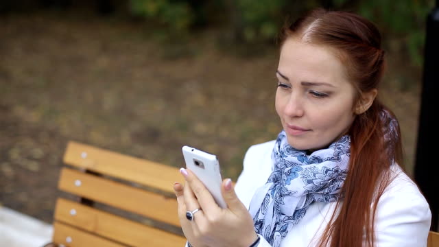 Rothaarige-Frau-mit-Smartphone-in-der-Hand-sitzt-auf-einer-Bank-im-Park-und-kommuniziert-in-sozialen-Netzwerken