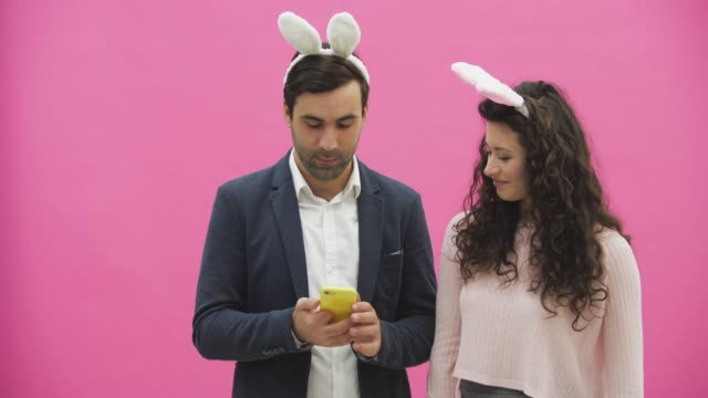 Young-lovers-couple-on-the-pink-background.-With-hackneyed-ears-on-the-head.-During-this,-they-make-a-photo-of-sephi-on-the-phone-showing-various-rabbit-movements-and-rejoice.