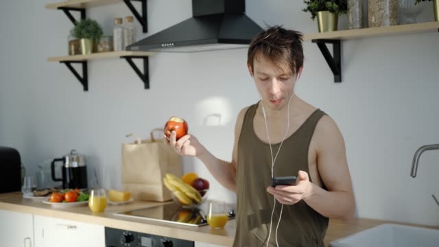 Fröhlicher-junger-Mann-isst-Apfel,-Musik-hören-in-leichter-Küche