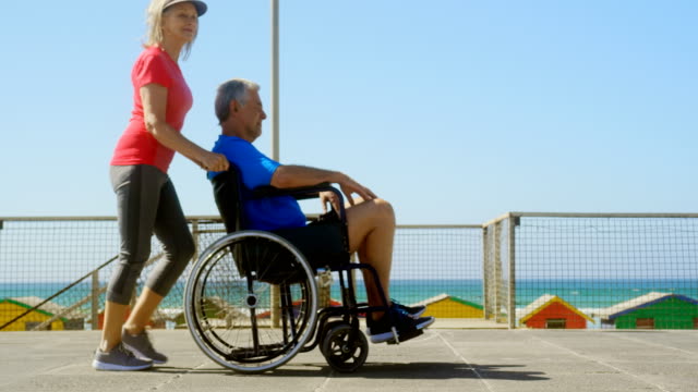 Seitenansicht-der-aktiven-Seniorkaukasschen,-die-ältere-Mann-auf-den-Rollstuhl-am-Strand-4k-drängt