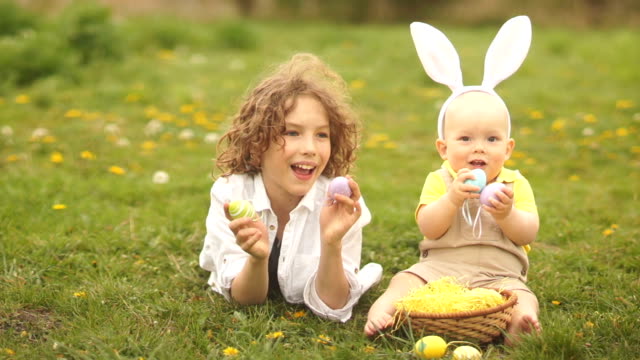 Two-brothers,-a-teenager-and-a-baby-are-resting-in-a-park-on-a-glade.-Easter-concept,-bunny-ears,-easter-basket-and-eggs.-A-dog-runs-past-them