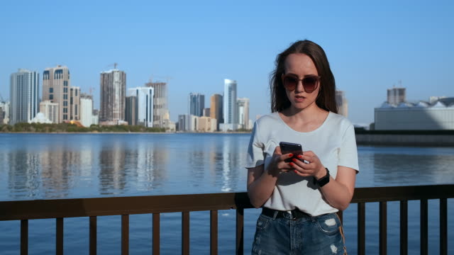 Hermosa-chica-con-el-pelo-largo-en-gafas-de-sol-usando-aplicación-de-teléfono-inteligente-en-Sunset-River-Quay-cerca-de-4K.