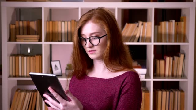 Retrato-de-primer-plano-de-joven-pelirroja-atractiva-estudiante-caucásica-en-gafas-usando-la-tableta-en-la-biblioteca-de-la-universidad