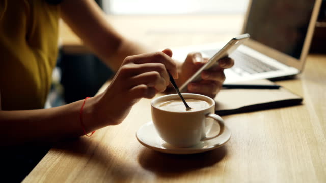 Primer-plano-de-las-manos-de-la-mujer-mezclando-café-en-taza-y-usando-teléfonos-inteligentes-relajándose-en-el-café