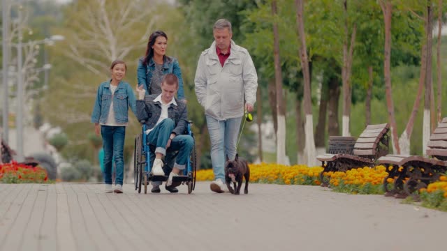 Eltern-gehen-den-Weg-mit-den-Kindern-und-dem-Hund.-Sohn-im-Rollstuhl-fährt-mit-seiner-Familie-die-Allee-entlang.