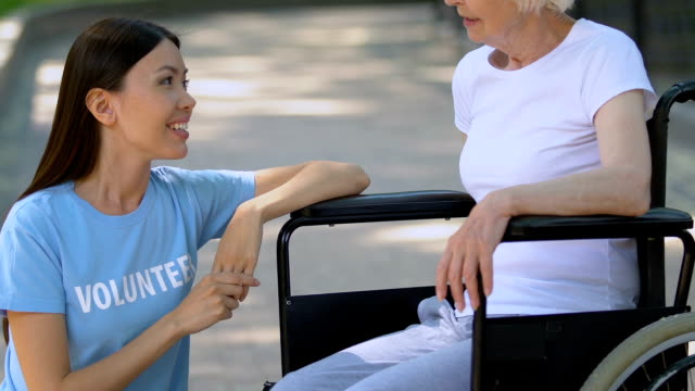 Voluntario-sonriente-hablando-con-una-vieja-paciente-en-silla-de-ruedas,-relájese-en-el-parque