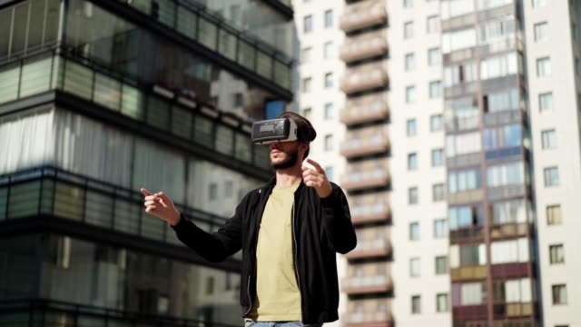 Tracking-slow-motion-shot-of-young-man-in-vr-glasses-standing-outdoors-in-city-street-and-exploring-virtual-reality