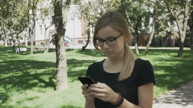 Perplexed-caucasian-woman-in-eyeglasses-walking-checking-smart-phone-content-and-then-stops-looking-at-camera-in-a-park