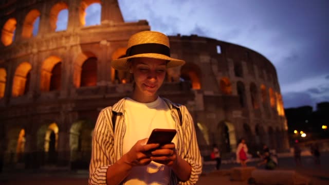 Fröhliche-weibliche-Touristin-im-Hut-zufrieden-mit-4G-Verbindung-in-Roaming-teilen-Multimedia-und-chatten-online-während-Sightseeing-in-Rom-in-der-Nähe-von-Kolosseum-am-Abend