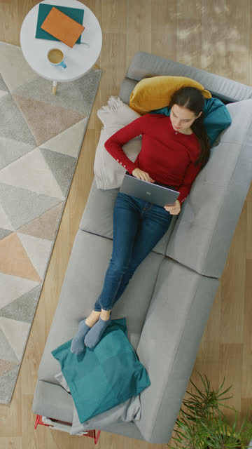 Chica-joven-en-blusa-roja-y-azul-jeans-está-acostado-en-un-sofá,-utilizando-un-ordenador-portátil.-Ella-mira-arriba-y-sonríe.-Vista-superior-con-Zoom.