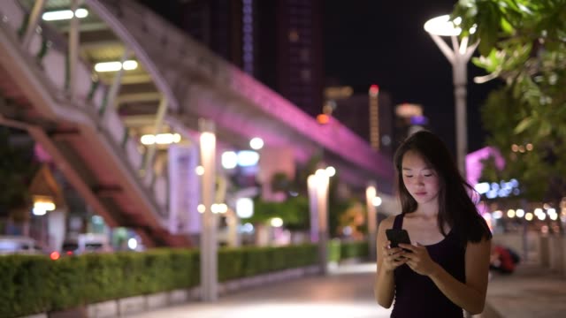 Junge-schöne-asiatische-Frau-mit-Telefon-in-der-Stadt-in-der-Nacht