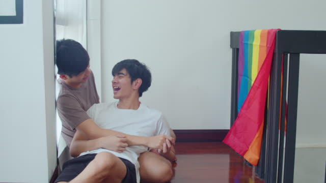 Asiatische-Homosexuell-Paar-liegen-und-umarmen-auf-dem-Boden-zu-Hause.-Junge-asiatische-LGBTQ+-Männer-küssen-glückliche-Entspannen-Ruhe-zusammen-verbringen-romantische-Zeit-im-Wohnzimmer-mit-Regenbogen-Flagge-im-modernen-Haus-am-Morgen.