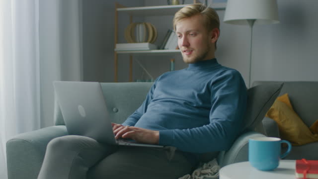 Porträt-von-Handsome-Blonde-Young-Man-Arbeitet-an-einem-Laptop-Computer,-während-auf-einem-Stuhl-in-seinem-gemütlichen-Wohnzimmer-sitzen.-Creative-Freelancer-entspannt-zu-Hause,-surft-im-Internet,-nutzt-Social-Media-und-entspannt