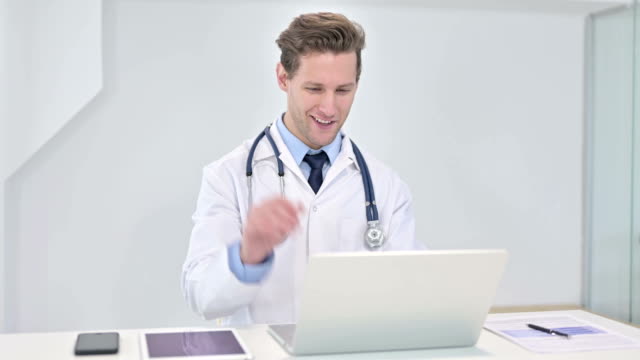 Fröhlicher-junger-Arzt-macht-Video-Chat-auf-Laptop-im-Büro