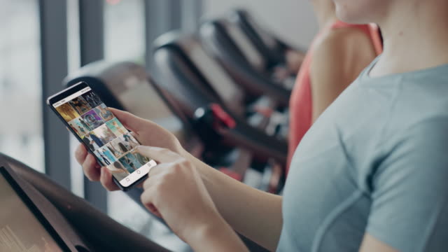 Close-up-of-a-Female-Using-Smartphone-in-a-Gym-on-a-Treadmill,-Browsing-Through-Motivational-Videos-on-Social-Network.-Finger-Tapping-Between-Videos-on-Social-App-Feed.-Mock-up-Application-Design.