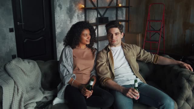 Man-and-woman-moved-to-new-flat.-They-smiling-and-enjoying-beer-while-sitting-on-gray-comfortable-couch-in-living-room-with-cardboard-boxes