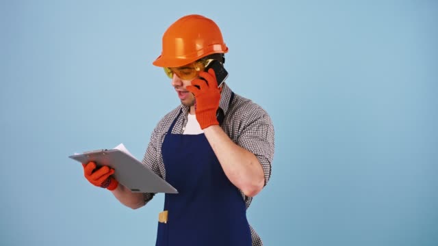 Young-man-in-safety-clothes,-hard-hat,-protective-goggles,-gloves.-Talking-by-smartphone,-looking-at-papers-on-clipboard.-Posing-on-blue-background