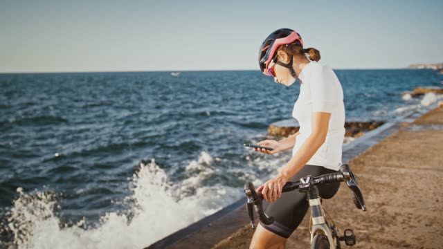 Girl-in-protective-helmet,-sunglasses-and-sportswear-is-sitting-on-bike-at-sea-embankment.-She-is-typing-on-her-cellphone.-Sunny-day,-slow-motion