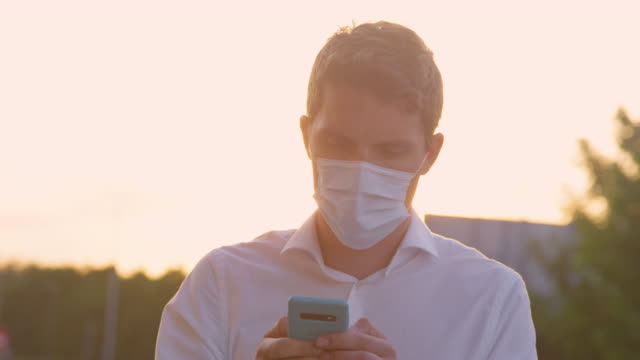 CLOSE-UP:-Young-man-wearing-a-medical-facemask-texts-on-a-sunny-summer-evening.