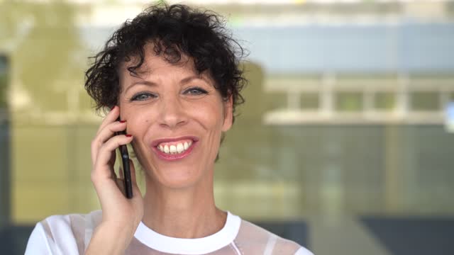 Zoom-in-close-up-shot-of-cheerful-mature-business-woman-talking-on-mobile-phone-outside-office