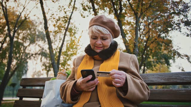 Rentnerin-betritt-eine-Reihe-ihrer-Kreditkarte-ins-Handy,-während-sie-im-Herbstpark-auf-der-Bank-sitzt