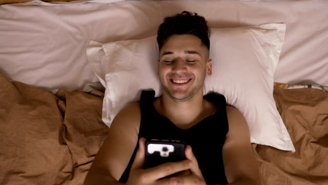 Smiling-young-man-networking-in-bed-before-sleeping-using-smartphone-on-social-media