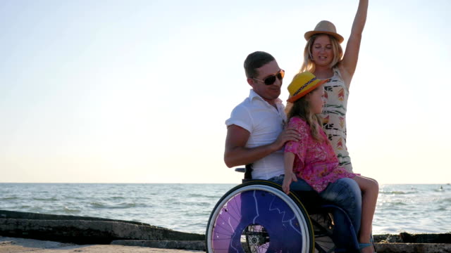 happy-invalid,-family-play-together-on-background-blue-sky,-girl-sitting-on-daddy-in-wheelchair-with-outstretched-arms