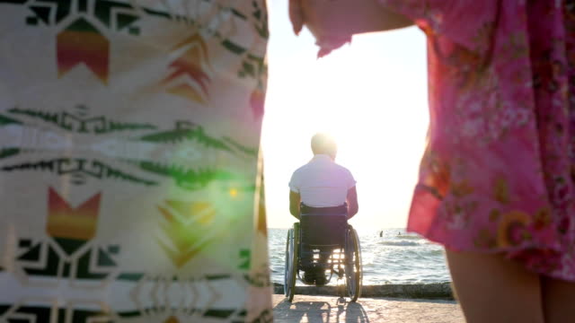 Disabled-man-in-wheelchair-with-family,-mother-and-daughter-holding-hands-in-background-father-sitting-in-wheelchairs-near-water