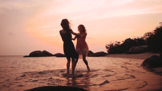 two-happy-dancing-girls-holding-hands-on-the-beach-at-sunset-in-slow-motion