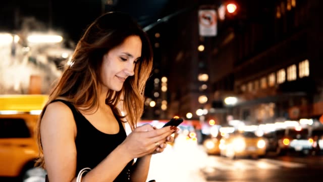 Portrait-of-young-happy-woman-standing-in-traffic-downtown-in-the-evening-in-New-York,-America-and-using-the-smartphone