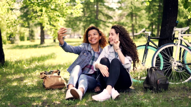 Hübsche-Mädchen-Freunde-nehmen-Selfie-Park-auf-Rasen-mit-Fahrrädern-im-Hintergrund-sitzen.-Gemischte-Rassen-Freundschaft,-moderne-Technologie-und-fröhliche-Menschen-Konzept.