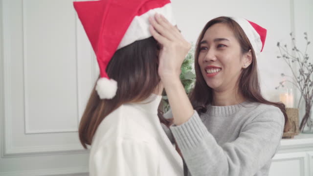 Asiatische-Lesben-Paar-geben-und-Weihnachtsmütze-miteinander-in-ihrem-Wohnzimmer-zu-Hause-im-Christmas-Festival-tragen.-Lebensstil-Lgbt-Frauen-glücklich-feiern-Weihnachten-und-Neujahr-Konzept.