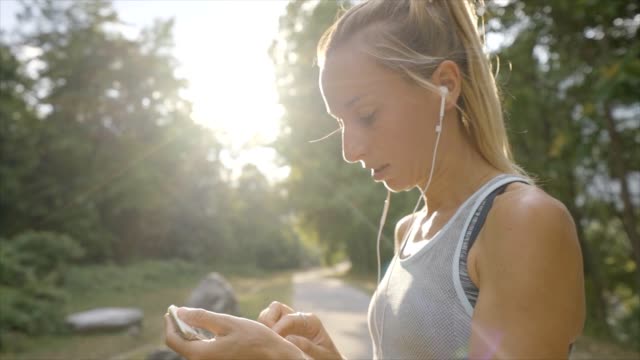 Young-woman-jogging-outdoors-using-mobile-phone-app-choosing-music-for-morning-training-in-forest-path.-People-healthy-lifestyle-concept---Slow-motion