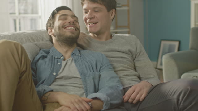 Retrato-de-una-linda-pareja-gay-masculina-en-el-hogar.-Se-sientan-en-un-sofá-y-mirar-a-la-cámara.-Pareja-abraza-a-su-amante-por-detrás.-Ellos-son-feliz-y-sonriente.-Habitación-tiene-un-Interior-moderno.