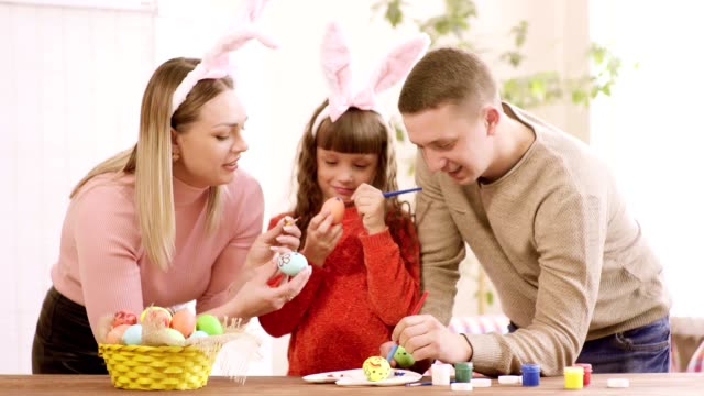 mamá,-papá-e-hija-apoyándose-en-la-mesa-decoran-los-huevos-de-Pascua.