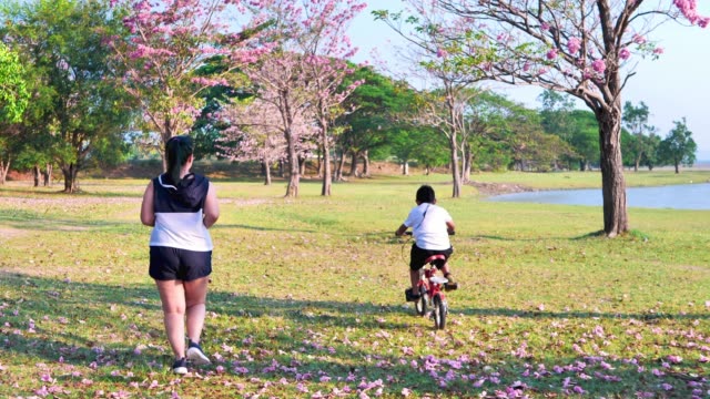 Una-mujer-asiática-gorda-corriendo-en-la-luz-del-sol-natural-por-la-noche,-junto-con-su-hijo-montando-en-bicicleta.--ejercicio-para-una-buena-salud.