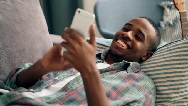 Hombre-alegre-usando-el-teléfono-inteligente-acostado-en-el-sofá-en-casa