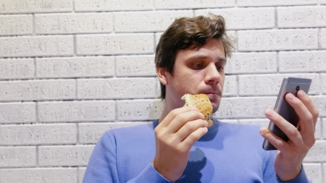 El-hombre-está-comiendo-una-hamburguesa-en-el-café-y-viendo-un-video-en-el-teléfono-móvil.