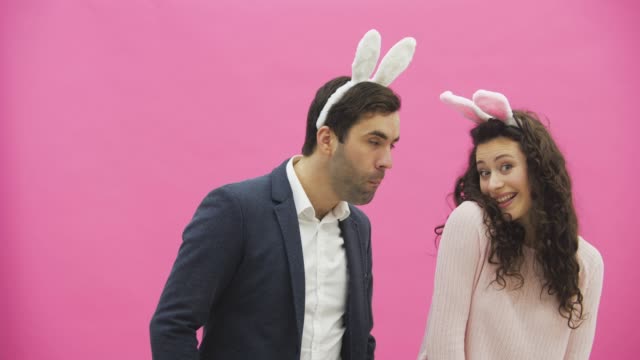 Beautiful-young-couple-standing-on-pink-background.-At-the-same-time,-the-girl-claps-her-husband,-he-in-turn-bites-her-hand.-Together-with-pink-barking-ears-on-the-head.-Happy-family-is-preparing-for-Easter.