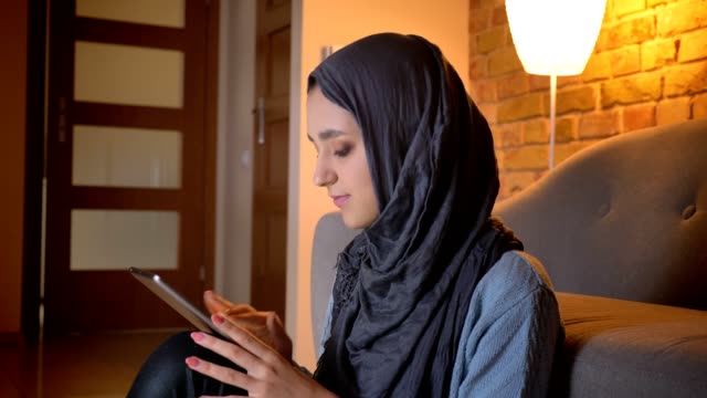 Closeup-shoot-of-young-attractive-muslim-female-in-hijab-typing-on-the-phone-while-sitting-on-the-floor-in-the-doorway-at-cozy-home