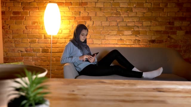 Tiro-de-primer-plano-de-la-joven-atractiva-de-la-web-femenina-de-surf-musulmana-en-el-teléfono-mientras-descansa-relajado-en-el-interior-de-la-acogedora-casa