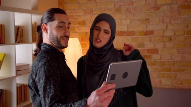Closeup-shoot-of-young-cheerful-muslim-couple-having-a-video-call-on-the-tablet-and-showing-off-their-newly-bought-apartment