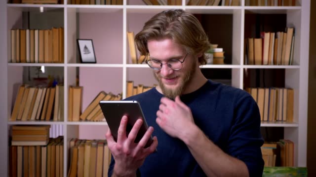 Closeup-shoot-of-adult-attractive-male-student-using-the-tablet-smiling-happily-in-the-university-library-indoors