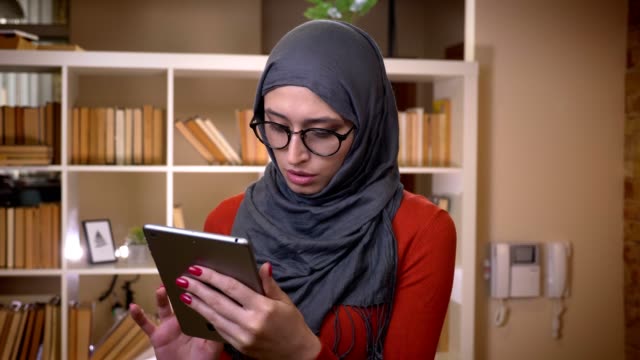Sesión-de-primer-plano-de-joven-atractiva-estudiante-musulmana-en-hiyab-navegando-en-la-tableta-de-pie-en-el-interior-de-la-universidad-de-la-biblioteca